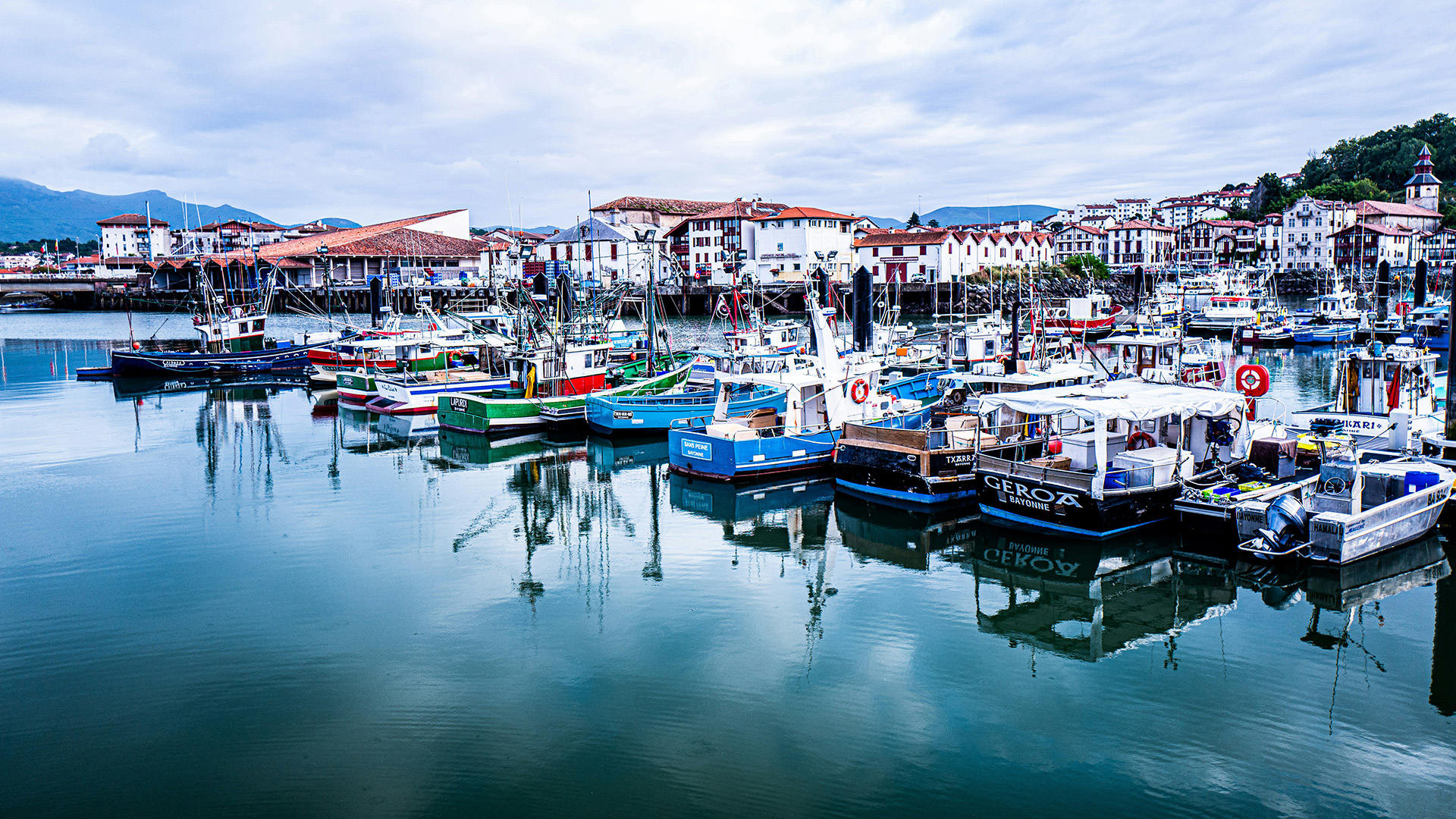 Colorful fishing boats reflected in a calm harbor below red-and-white Basque houses